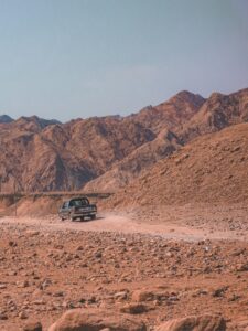 A rugged off-road vehicle kicks up dust as it travels through a vast desert landscape, with rolling hills of reddish-brown rock in the background