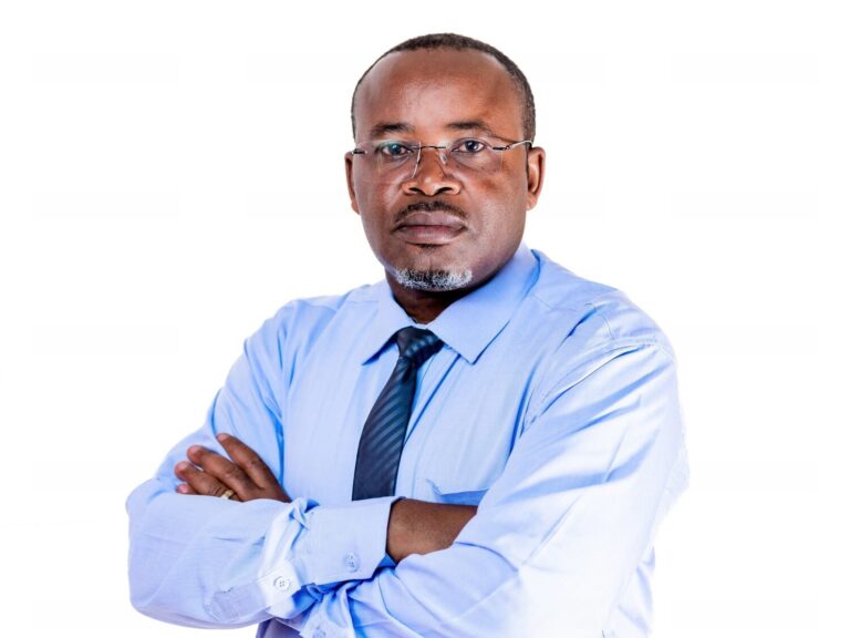 Dr De-Joseph Kakisingi, a humanitarian leader from the Democratic Republic of Congo, looks straight at the camera. He is wearing a blue shirt and tie, and is standing with his arms crossed against a white background.