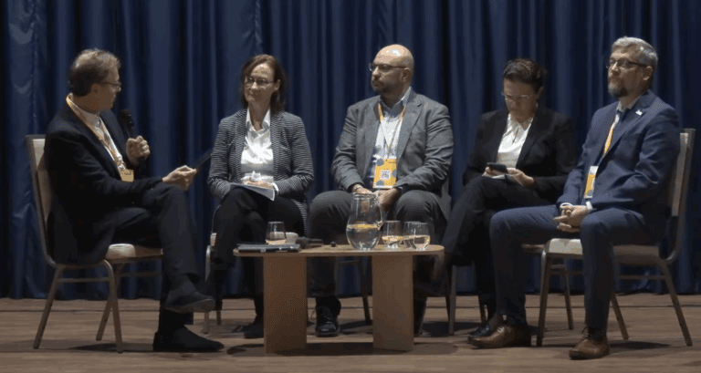 Floris Faber moderates a panel discussion at the Ambrela Development Forum in Bratislava. Four speakers sit on stage beside him, listening attentively. A small table with water glasses and microphones is in front of them, with dark blue curtains forming the backdrop.